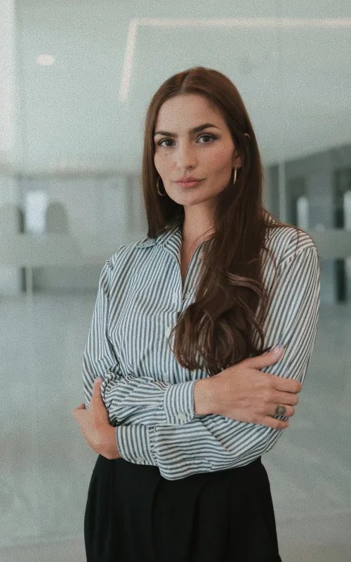 A woman with long brown hair in a striped shirt, arms folded, in a bright glass-walled interior