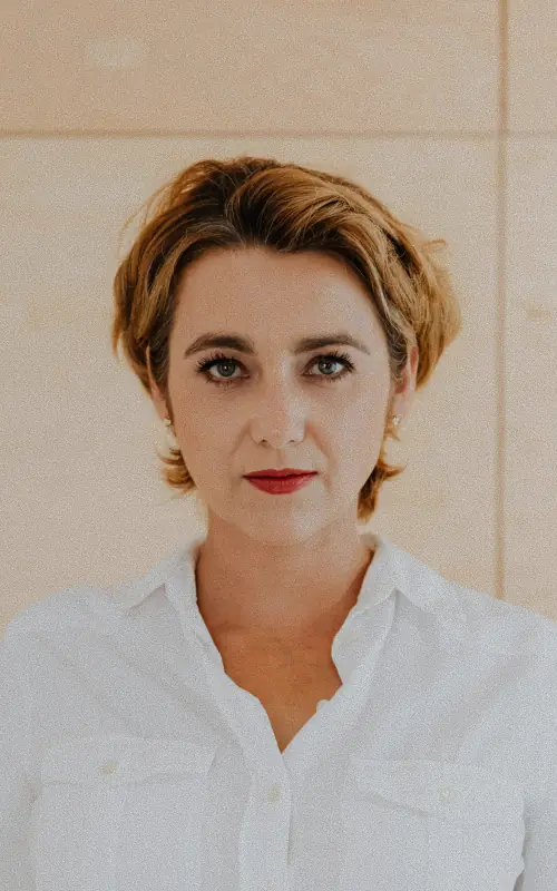 A woman with short strawberry blonde hair and red lipstick in a white shirt against a beige tiled wall