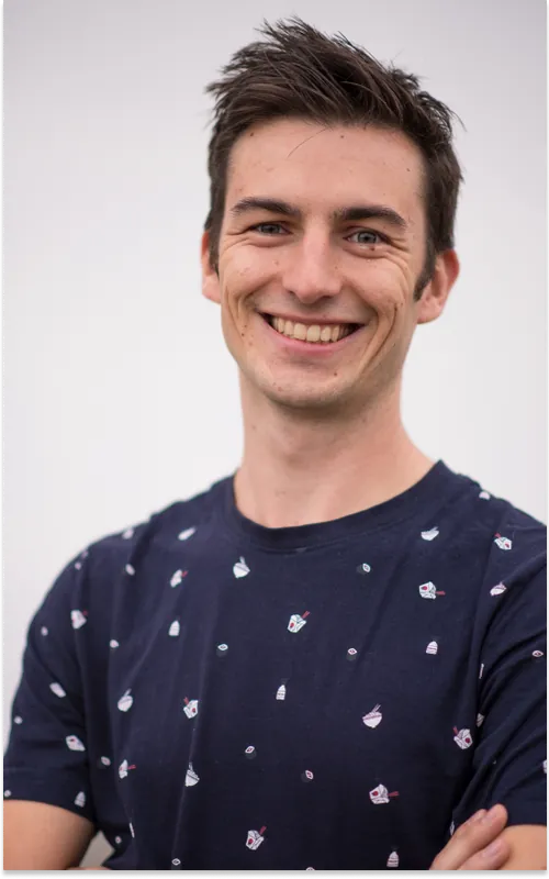 A smiling young man in a navy t-shirt printed with small food icons, arms folded, against a pale grey backdrop