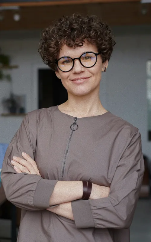 A woman with short curly brown hair and round glasses in a taupe zip-neck top, arms folded, in a warm interior